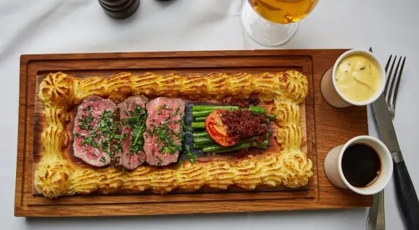 Sliced steak with green beans, tomato, and mashed potato border on a wooden board, with beer and sauces.