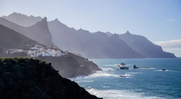 En vit by klättrar på en grön sluttning vid havet, med dramatiska berg i bakgrunden. En dröm om flygtid Teneriffa.