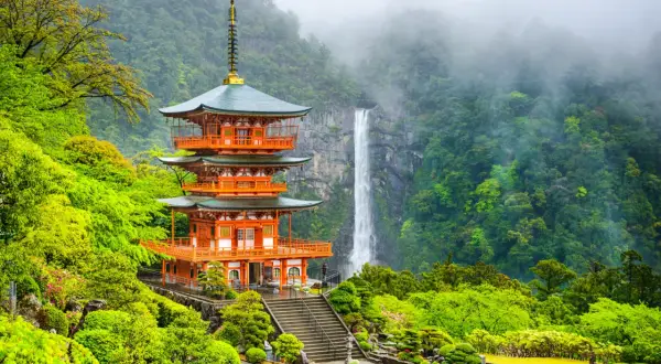 En orange pagod med gröna tak står vid en forsande vattenfall i en frodig, grön miljö. En del av Kumano Kodo-leden.