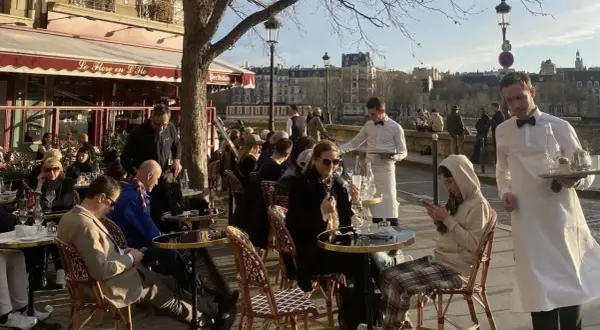 En solig dag på en restaurang i Paris. Människor sitter vid borden utomhus, och servitörer bär brickor med mat och dryck.