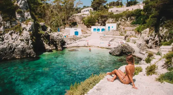 En kvinna sitter på en klippa vid en turkos vik i Polignano a Mare. Vita byggnader med blå dörrar syns på stranden.