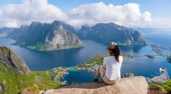 En person sitter på en klippa och blickar ut över en dramatisk kustlinje med berg och en by. En fantastisk vy för lofoten vandring.