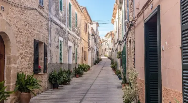 En smal, stenlagd gata i Alcudia old town kantas av gamla hus med gröna fönsterluckor och krukväxter.