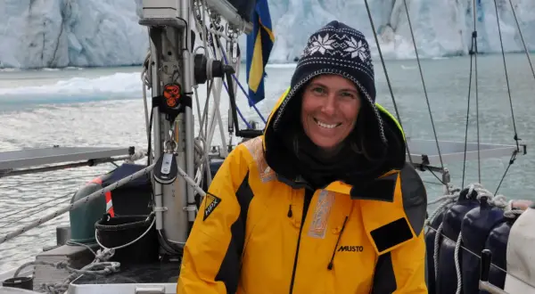 Glad kvinna i gul jacka på segelbåt framför glaciär. Drömmen att segla jorden runt är nära.