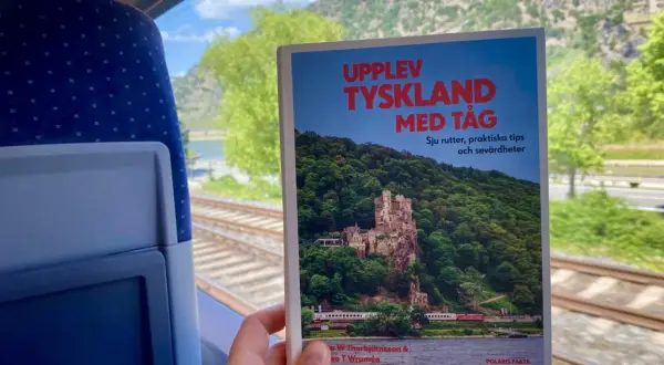 En hand håller en bok om att uppleva Tyskland med tåg, med en slående bild av ett slott vid en flod. Utsikten från tågfönstret visar räls och gröna kullar.