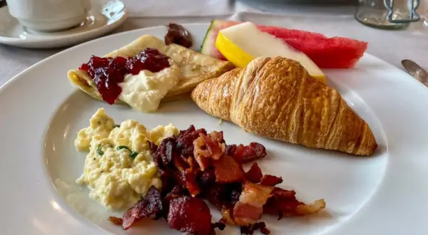 En härlig **continental frukost** med krämiga äggröror, knaprigt bacon, en croissant, pannkakor med sylt och grädde, samt färsk frukt.