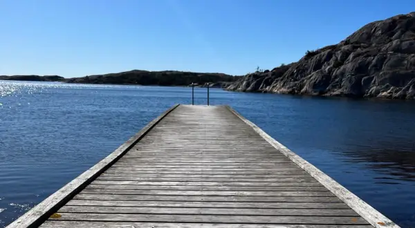 Träbrygga leder ut mot det glittrande vattnet vid Grebbestad strand. Klippor och öar i bakgrunden under en klarblå himmel.