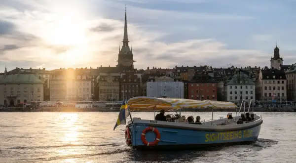 A blue sightseeing boat with a yellow striped canopy sails on water in Stockholm, with historic buildings and a church spire in the background at sunset.