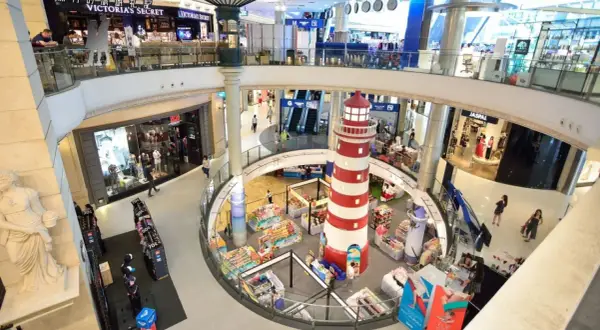 Inne i Terminal 21 Bangkok, ett shoppingcenter med flera våningar, syns en stor rödvit fyr i mitten. Folk shoppar och rör sig mellan butiker som Victoria's Secret.