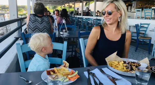 Familj njuter av en måltid på Costas Kitchen Kallithea. En kvinna ler, en pojke äter, och maten ser utsökt ut.