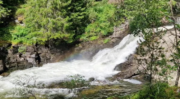 Vattenfall i Idre, med en regnbåge som skimrar i dimman från det forsande vattnet. Omges av grönskande träd och klippor.