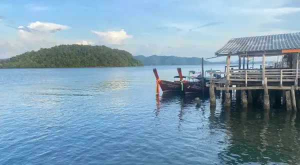 Två longtailbåtar vid en träbrygga på Koh Jum. En grönskande ö syns i bakgrunden under en molnig himmel.