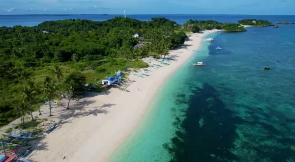 En vit sandstrand med palmer och traditionella filippinska båtar. Vattnet är turkost och klart, med en grönskande ö i bakgrunden. En idyllisk vy från Cebu, Filippinerna.