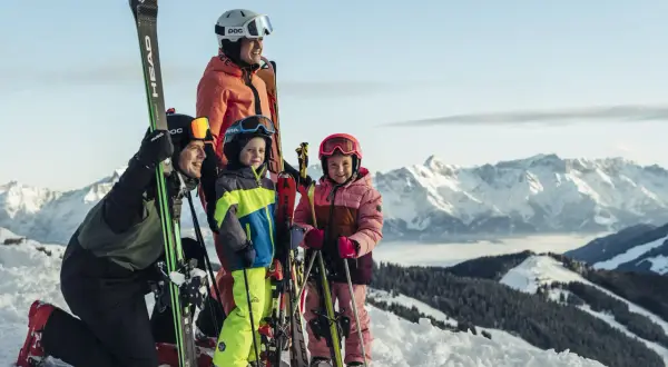 En glad familj njuter av sin skidresa i Alperna. Barnen ler mot kameran med skidor och stavar, redo för äventyr i snön.