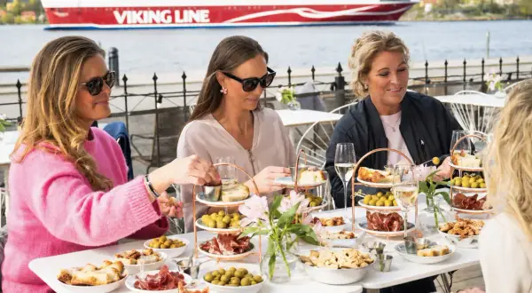 Kvinnor njuter av mat och dryck vid ett bord på en sq restaurang med Viking Line-färja i bakgrunden.
