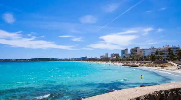 En livlig Cala Millor Beach med turkost vatten, solbadande människor och hotell längs kusten under en klarblå himmel.