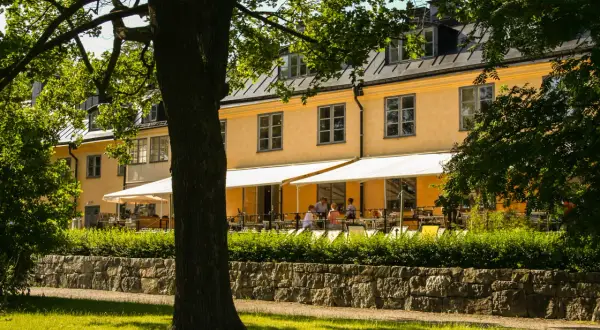 En gul byggnad med vita markiser och uteservering. En lång rad med träd skymmer delvis vyn.