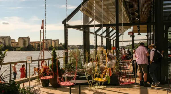 Sommar vid piren Hornsberg med folk som njuter av utsikten över vattnet och byggnader. Färggranna gungor och sittplatser utomhus.