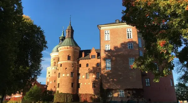 A red brick medieval castle with green domed towers under a clear blue sky, framed by autumn trees.
