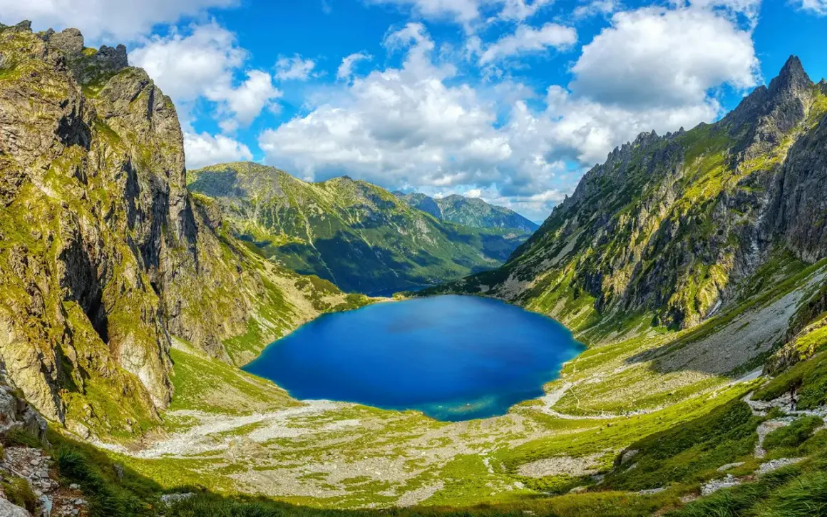 Blågrönt Morskie Oko, en sjö omgiven av gröna bergssluttningar och dramatiska klippor under en molnig himmel.
