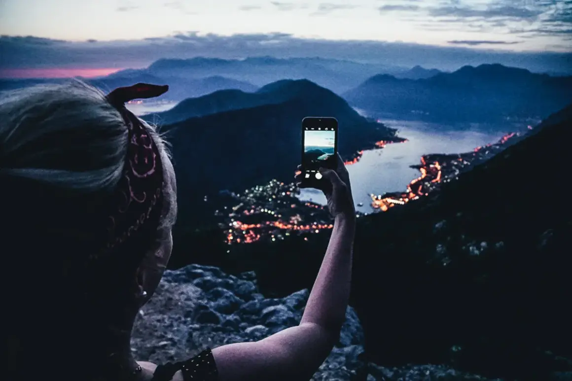 En person tar en bild med mobilen av en stad vid havet i Europa, med berg i bakgrunden.