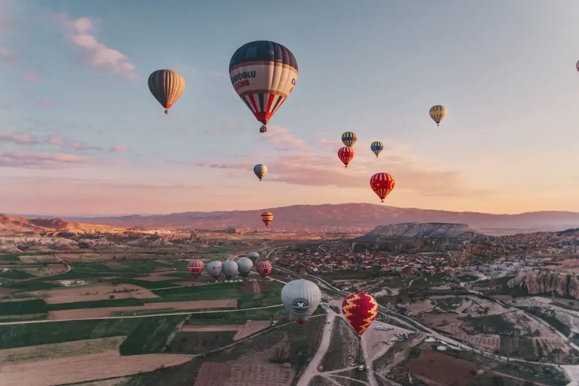 En magisk luftballong tur i Turkiet vid soluppgången. Många färgglada ballonger svävar över ett böljande landskap med byar och klippformationer.