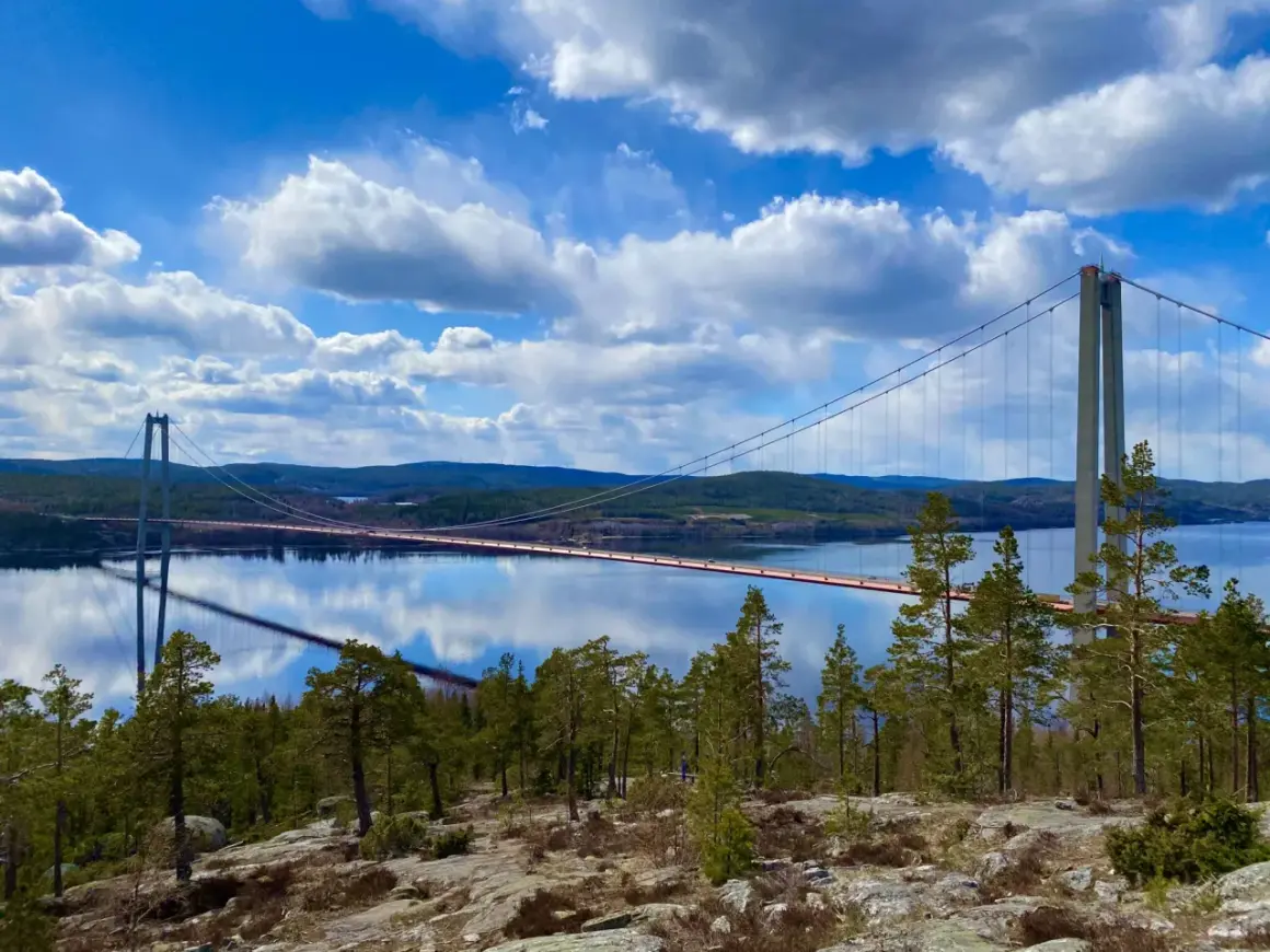 A suspension bridge spans a calm body of water under a bright blue sky with fluffy white clouds.