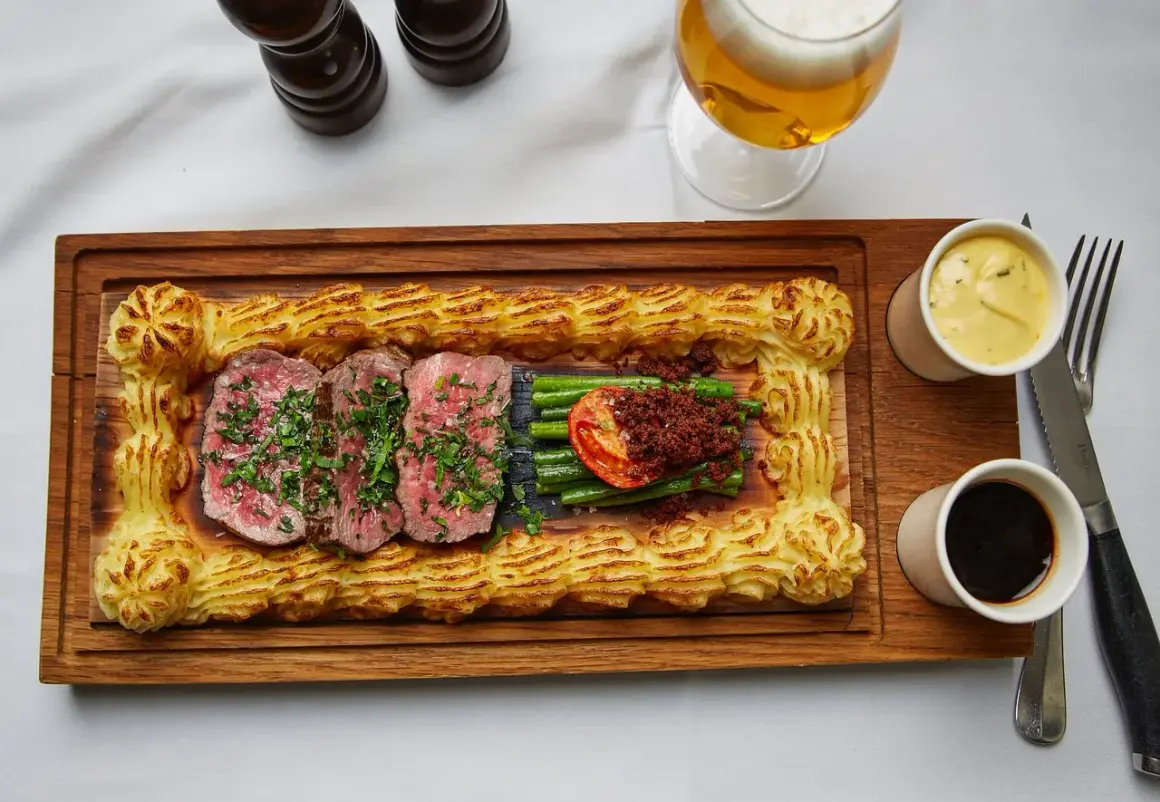 Sliced steak with green beans, tomato, and mashed potato border on a wooden board, with beer and sauces.