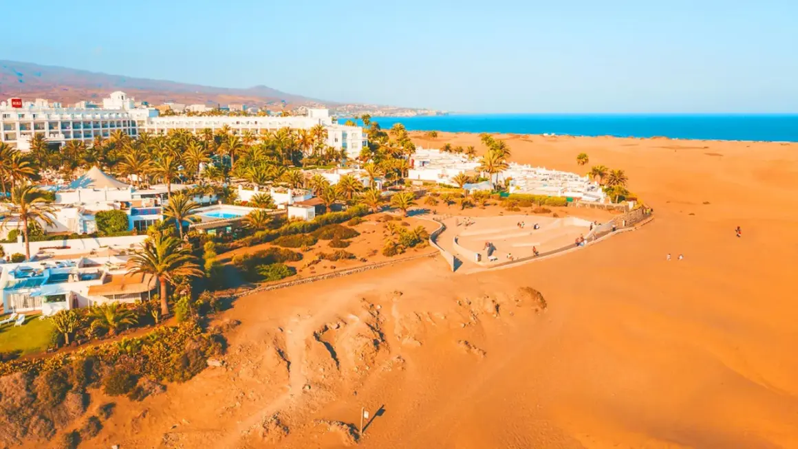 Vacker vy över Dunas de Maspalomas med vita hotell, palmer och gyllene sanddyner mot det blå havet.