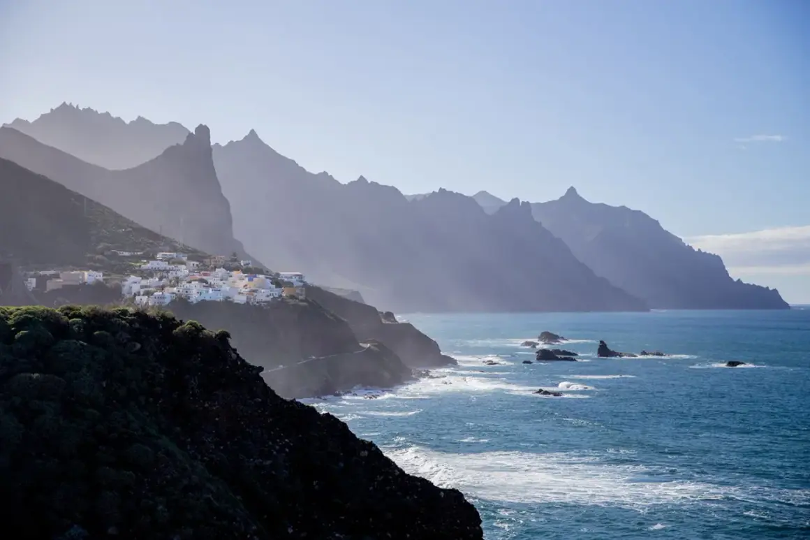 En vit by klättrar på en grön sluttning vid havet, med dramatiska berg i bakgrunden. En dröm om flygtid Teneriffa.