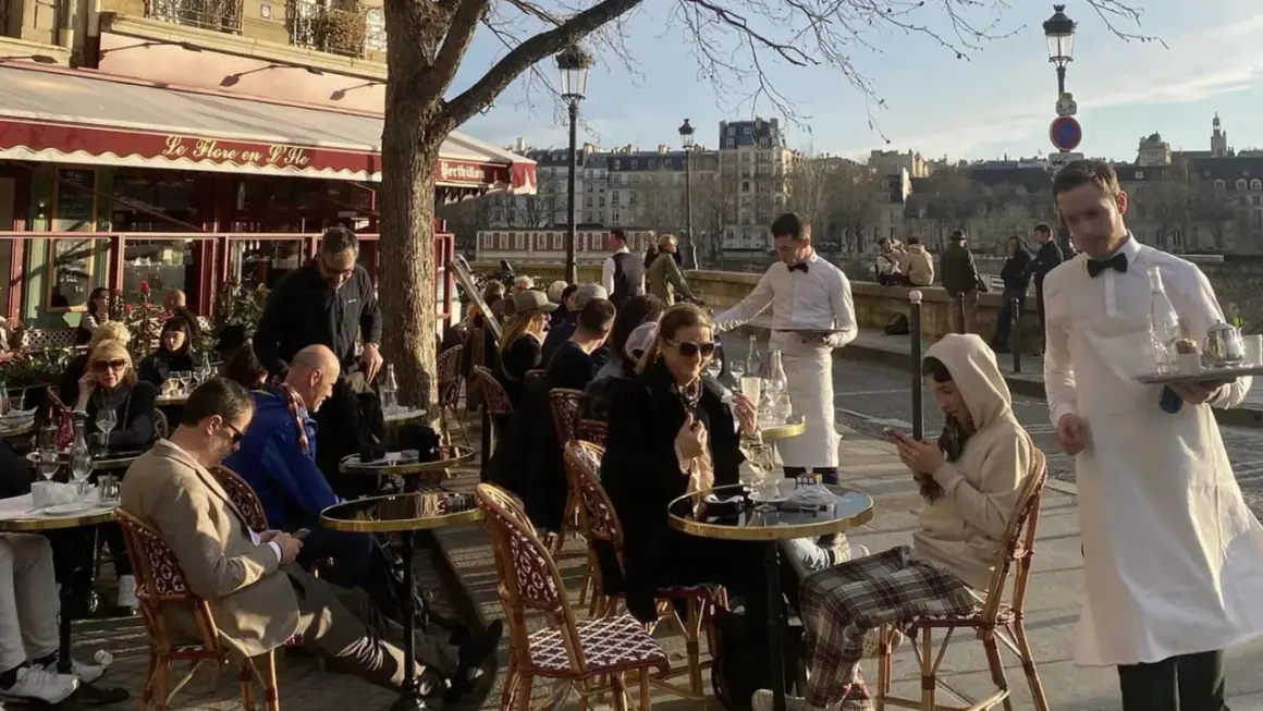 En solig dag på en restaurang i Paris. Människor sitter vid borden utomhus, och servitörer bär brickor med mat och dryck.