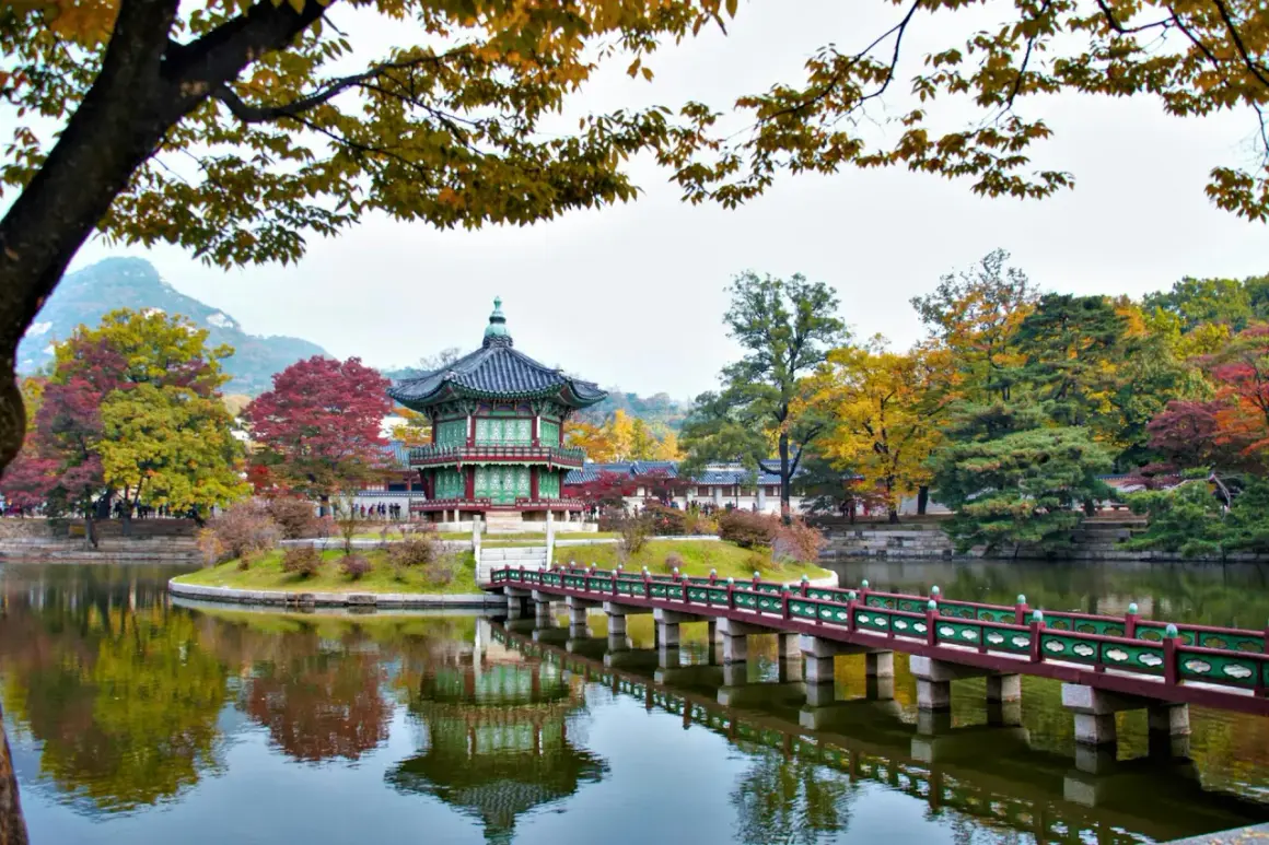 En vacker höstbild från Seoul, Sydkorea. Ett traditionellt koreanskt pagodliknande hus står på en ö i en damm, med en bro som leder dit. Träden runt omkring har höstfärger.