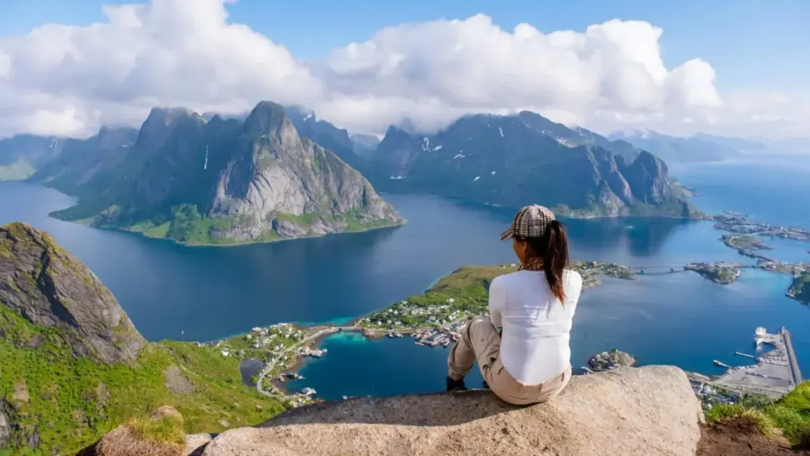 En person sitter på en klippa och blickar ut över en dramatisk kustlinje med berg och en by. En fantastisk vy för lofoten vandring.