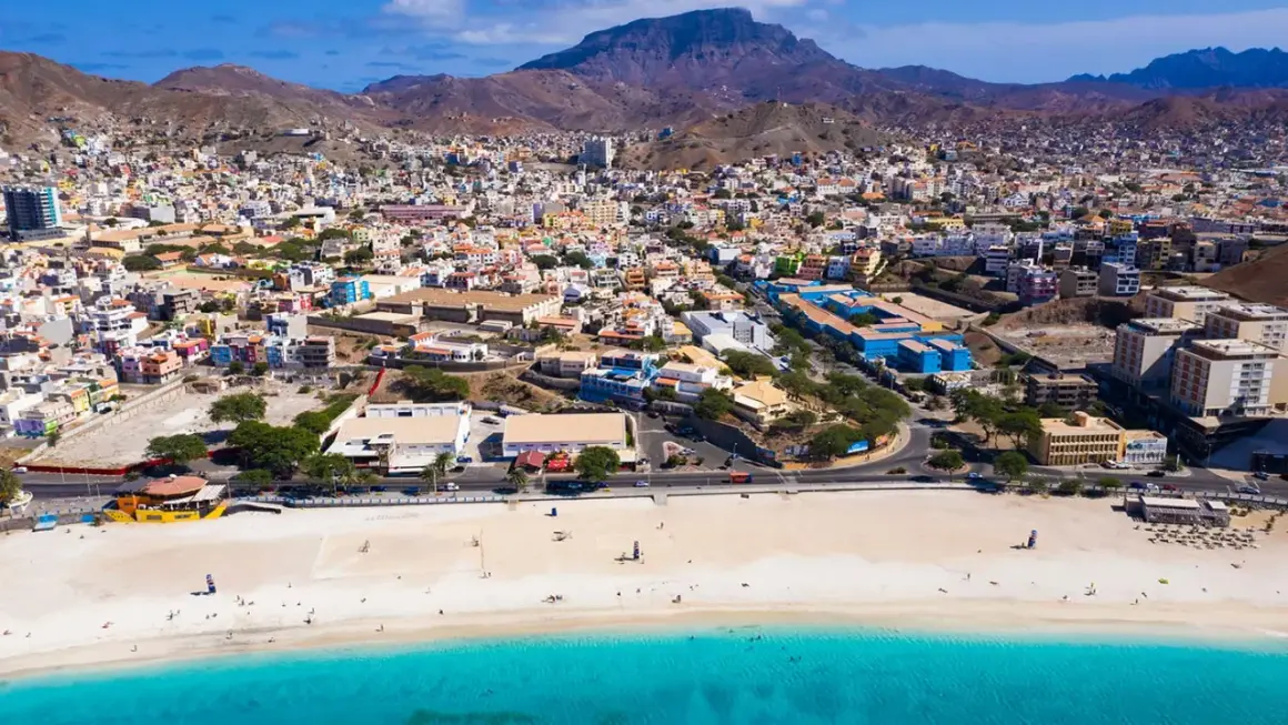 Färgstark stad i Kap Verde med en strand och turkost vatten. Berg reser sig bakom den tätbebyggda staden.