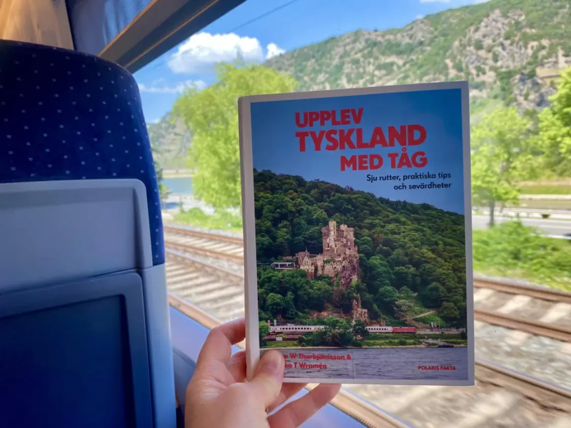 En hand håller en bok om att uppleva Tyskland med tåg, med en slående bild av ett slott vid en flod. Utsikten från tågfönstret visar räls och gröna kullar.
