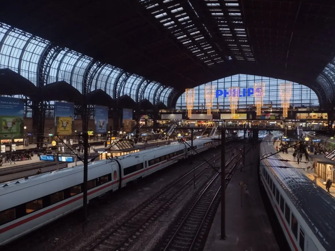 Tågstation med flera spår och vita tåg. En stor glas- och stålkonstruktion täcker taket. Philips-logotypen lyser i blått. Det är ett sj tåg till Hamburg.