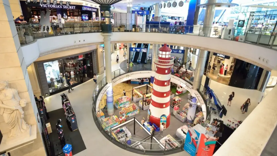 Inne i Terminal 21 Bangkok, ett shoppingcenter med flera våningar, syns en stor rödvit fyr i mitten. Folk shoppar och rör sig mellan butiker som Victoria's Secret.