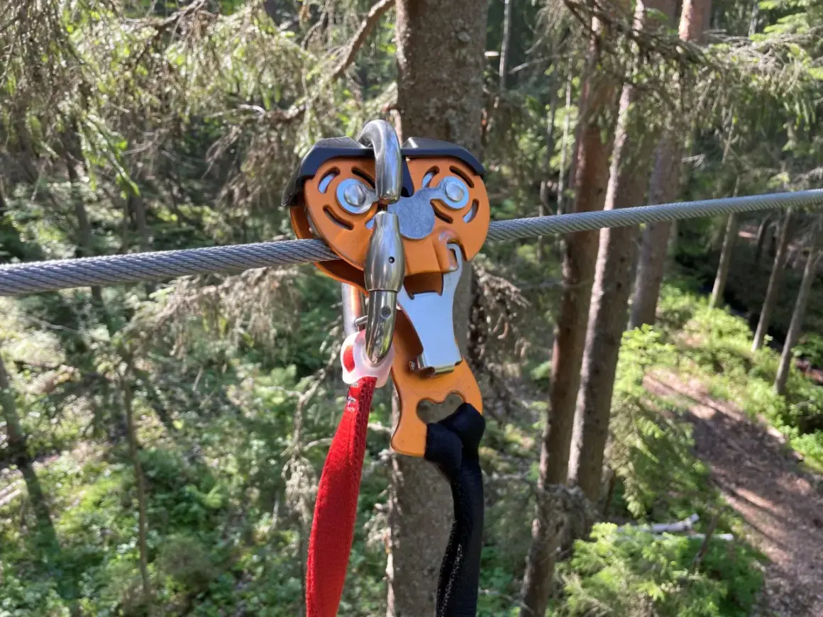 Nära en zipline i Umeå, en orange vajerklämma med röda och svarta band hänger redo för äventyr i skogen.