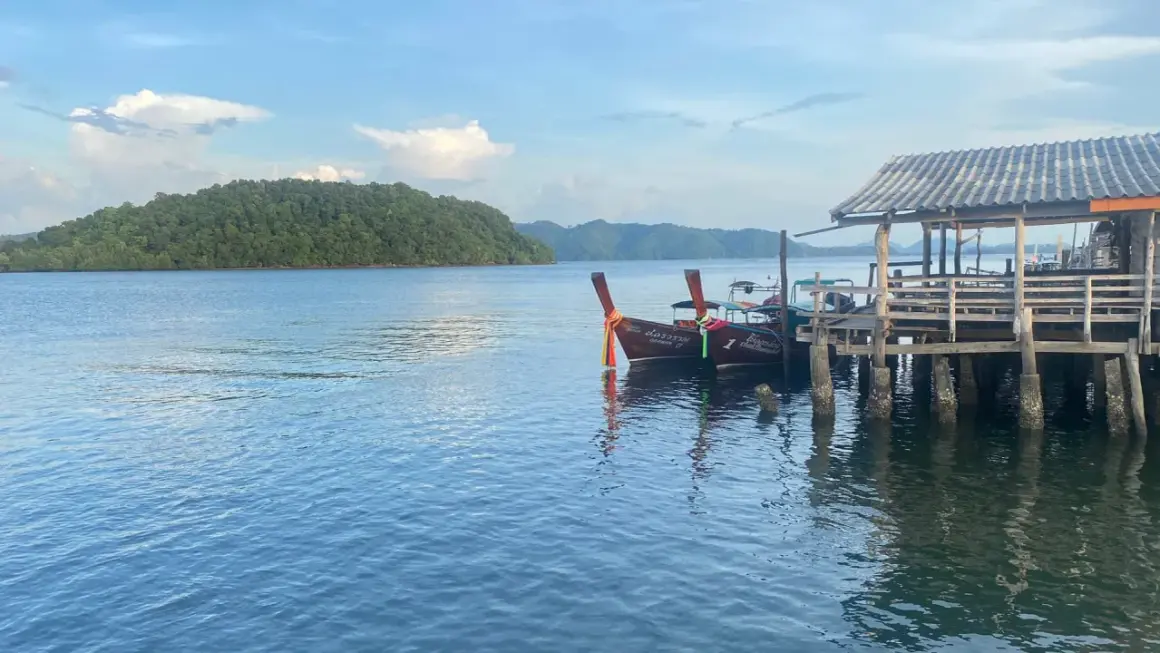 Två longtailbåtar vid en träbrygga på Koh Jum. En grönskande ö syns i bakgrunden under en molnig himmel.
