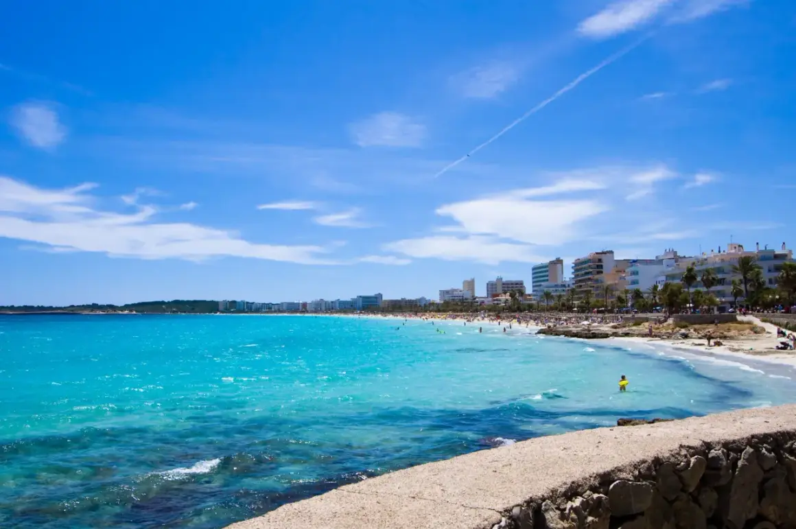 En livlig Cala Millor Beach med turkost vatten, solbadande människor och hotell längs kusten under en klarblå himmel.