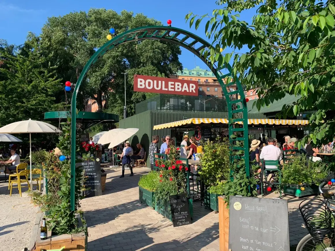 Sommar vid boulebar Rålambshov. Folk sitter vid bord under gula parasoller, grönska och röda blommor.