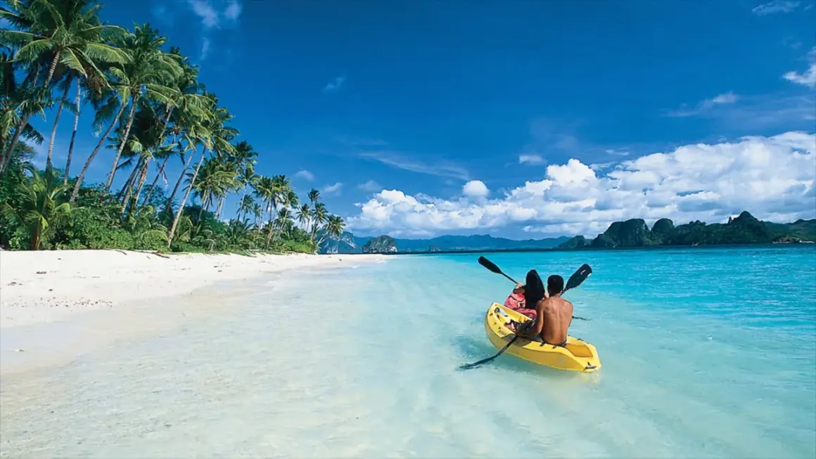 Två personer paddlar kajak i det turkosa vattnet vid en vit sandstrand med palmer på Palawan, Filippinerna.