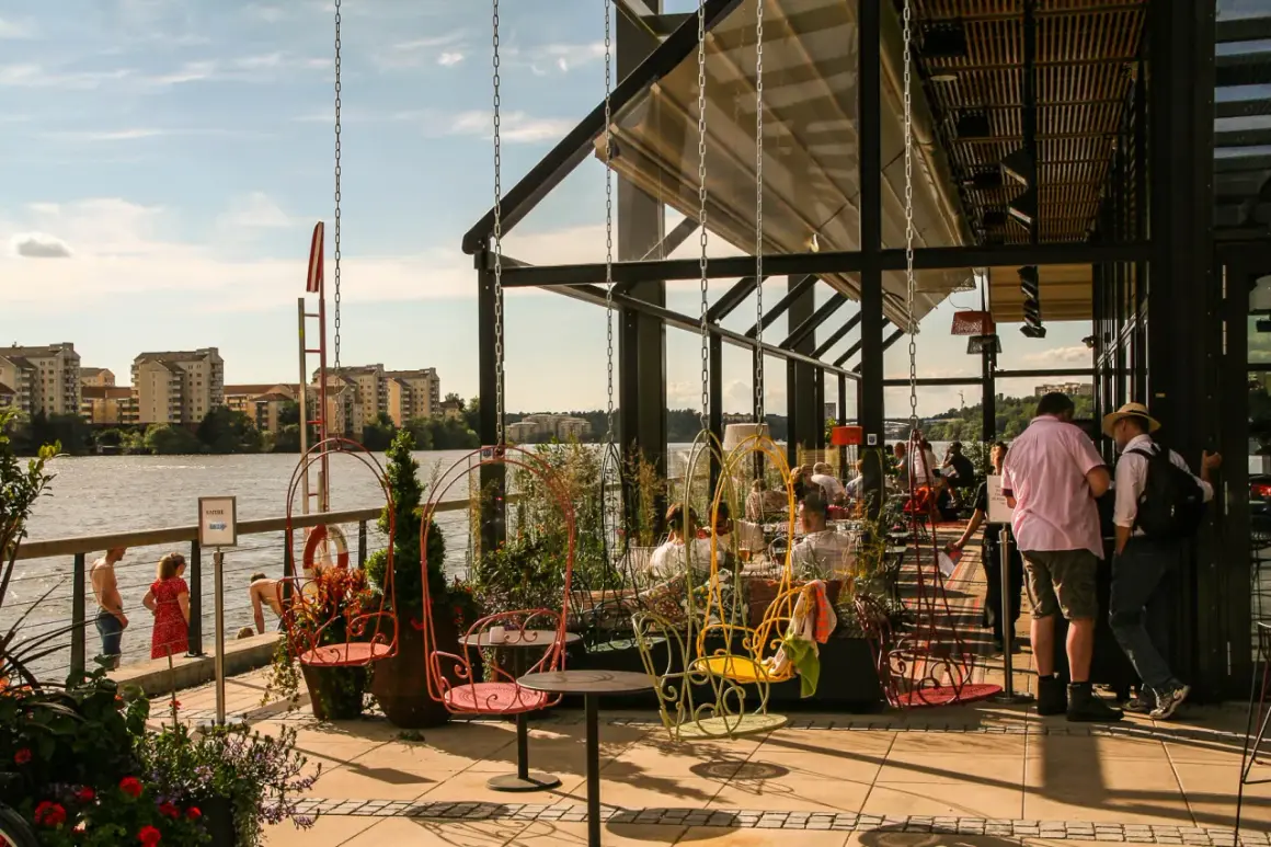 Sommar vid piren Hornsberg med folk som njuter av utsikten över vattnet och byggnader. Färggranna gungor och sittplatser utomhus.