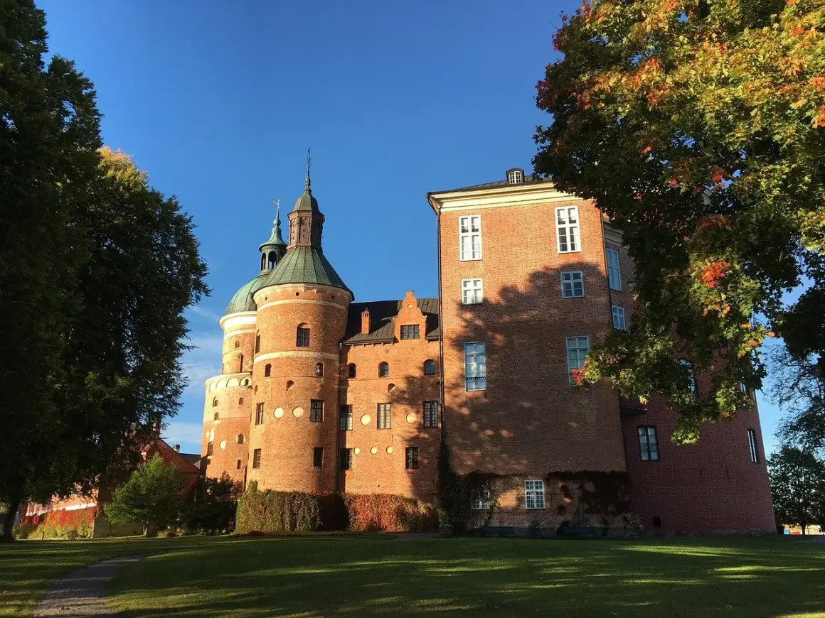 A red brick medieval castle with green domed towers under a clear blue sky, framed by autumn trees.