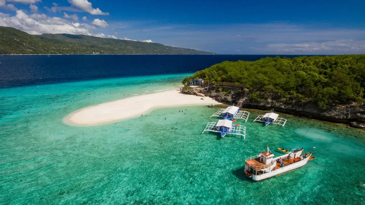 Vacker sandbank och turkost vatten vid Cebu, Filippinerna. Båtar guppar i det klara vattnet, en grönskande ö i bakgrunden.