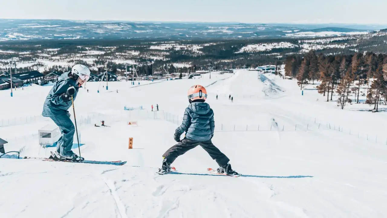 Tv&aring; barn njuter av en solig dag i skidbackarna i Dalarna. En vuxen l&auml;r ett barn &aring;ka skidor.