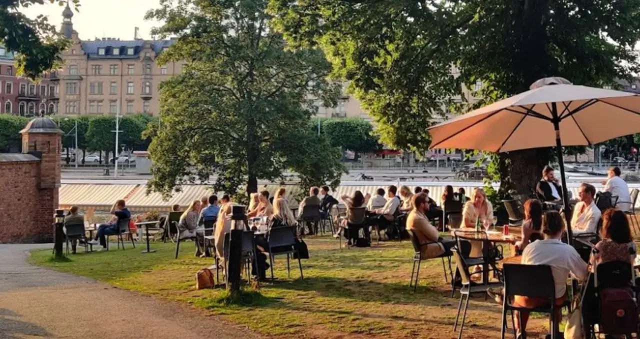 M&auml;nniskor njuter av en solig dag p&aring; en sommar restaurang i Stockholm, med utsikt &ouml;ver vattnet och vackra byggnader.