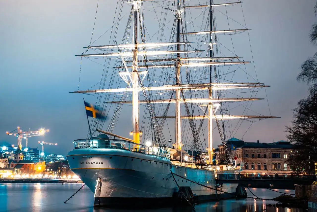 En ståtlig skonare, AF Chapman, ligger vid kajen. En lång rad av ljus lyser upp Skeppsholmen i bakgrunden.