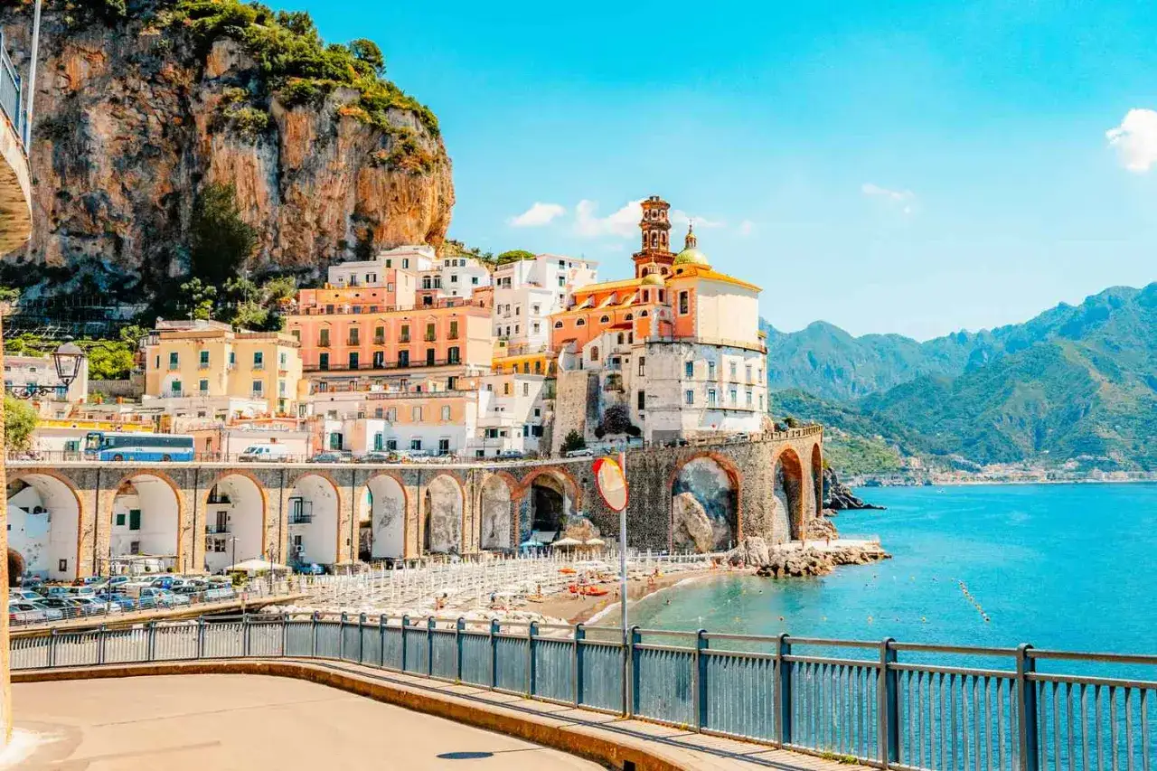 Färgstarka hus klättrar uppför klippan vid havet, ett dröm italien resmål. En strand med parasoller och en kyrka med kupol.