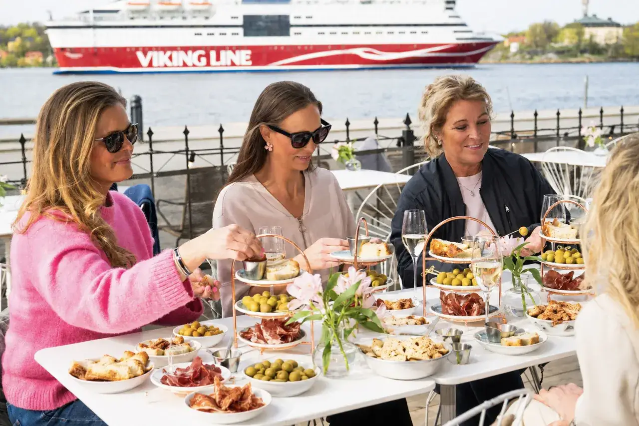 Kvinnor njuter av mat och dryck vid ett bord på en sq restaurang med Viking Line-färja i bakgrunden.